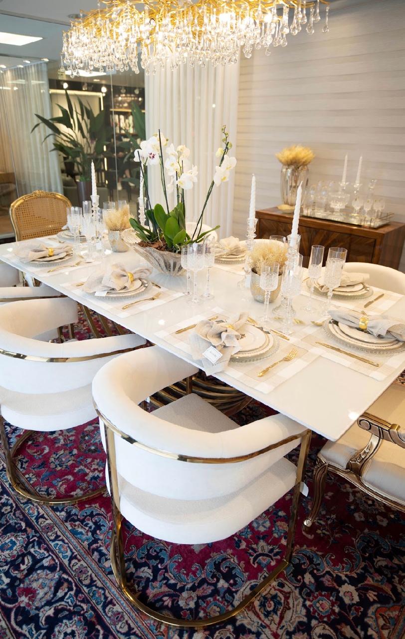Mesa de jantar posta com cadeiras brancas de base dourada, lustre de cristais e orquídeas