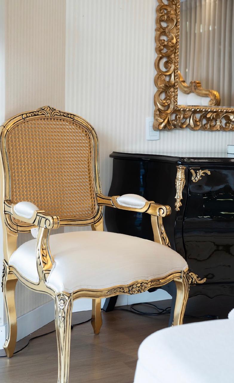 Poltrona clássica dourada com encosto de palhinha ao lado de cômoda preta laqueada
