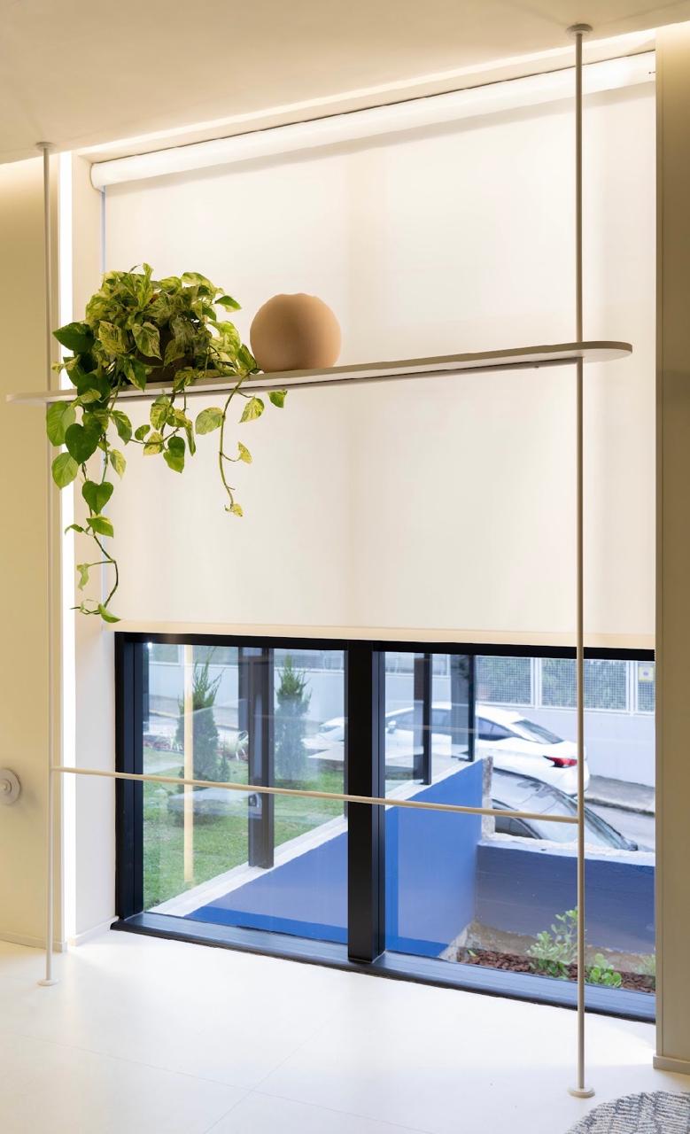Persiana rolô clara instalada em vão de vidro com prateleira e planta pendente