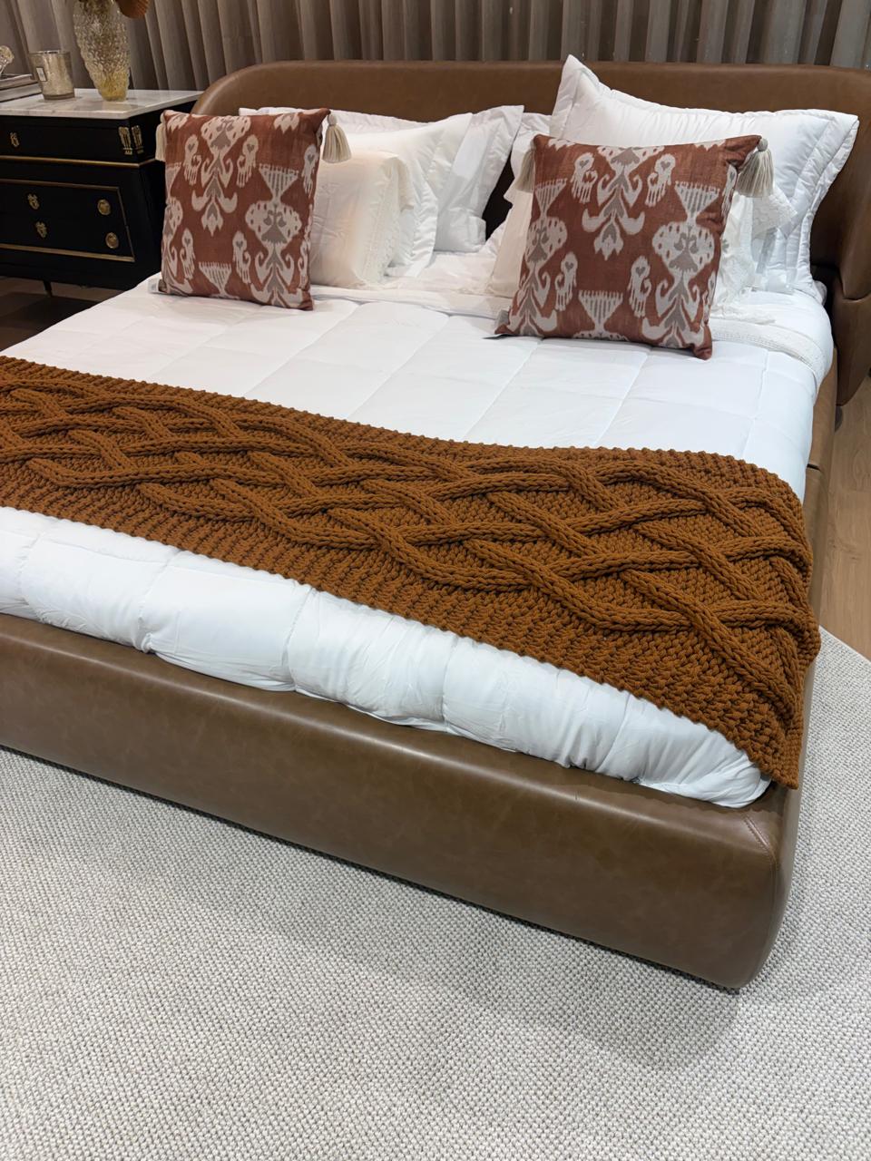 Cama estofada em couro caramelo com roupa de cama branca, manta de tricô caramelo e almofadas estampadas